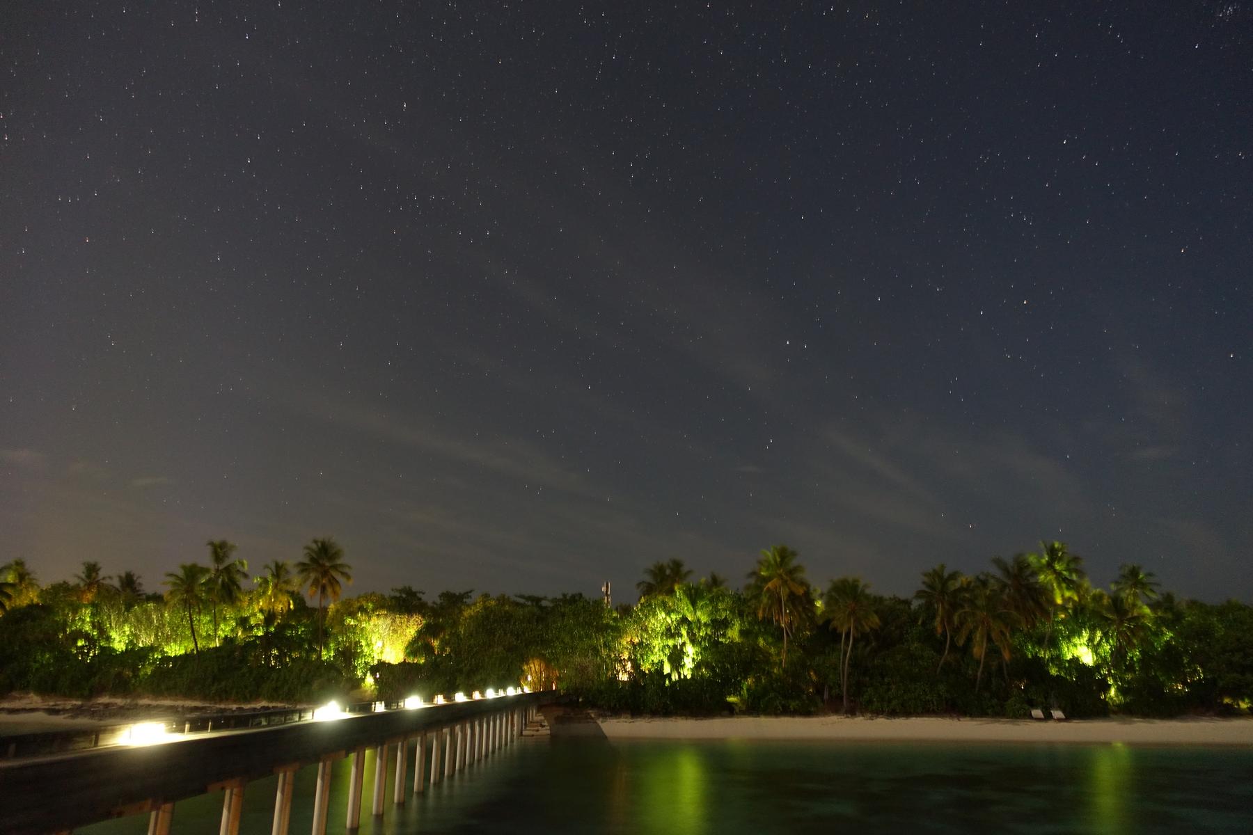 A starry night sky over a lit pier leading to an illuminated palm-covered island.