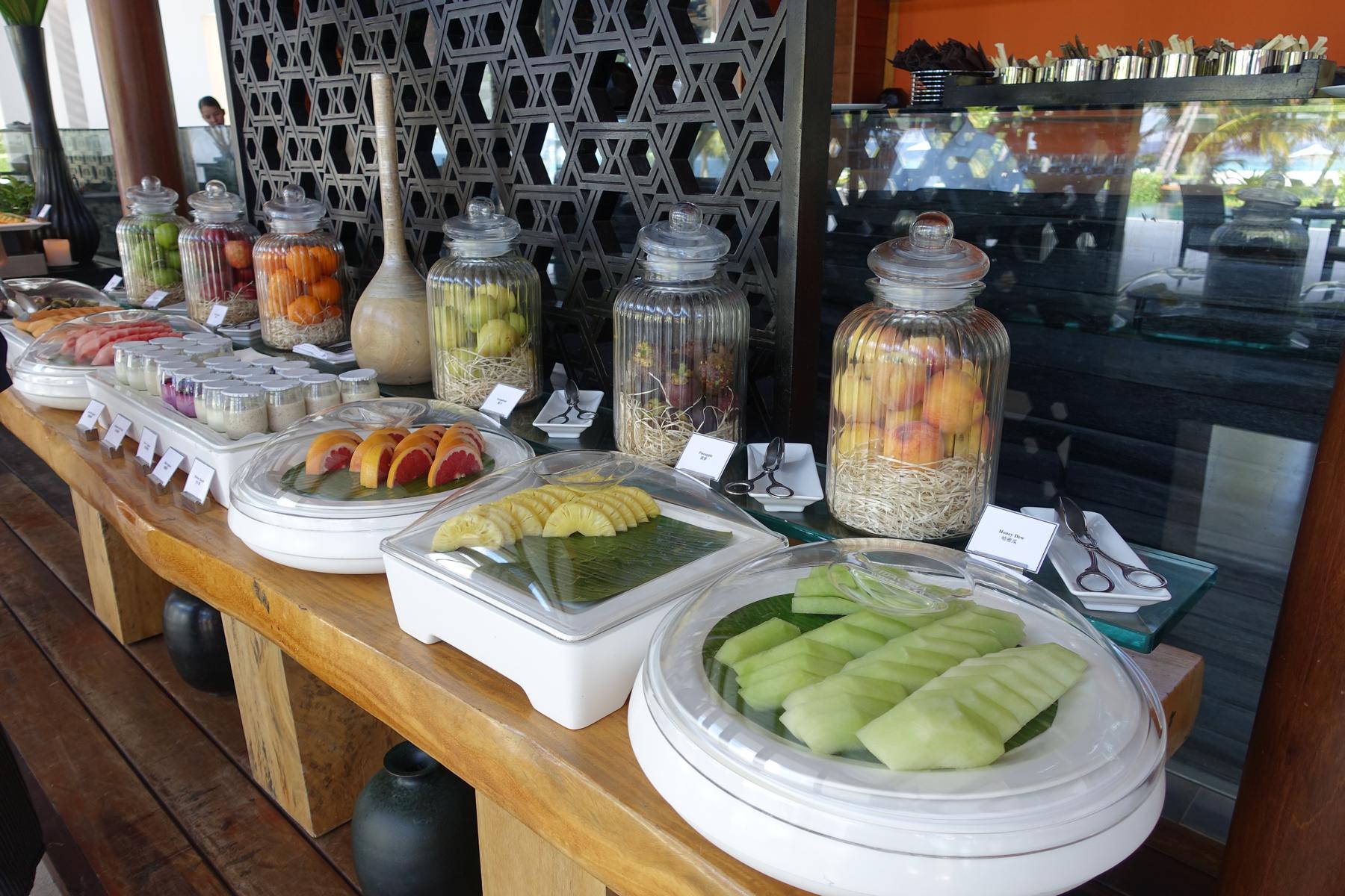 An elegant breakfast buffet displays whole fruits in decorative glass jars, various sliced fresh fruits in covered dishes, and individual yogurts.