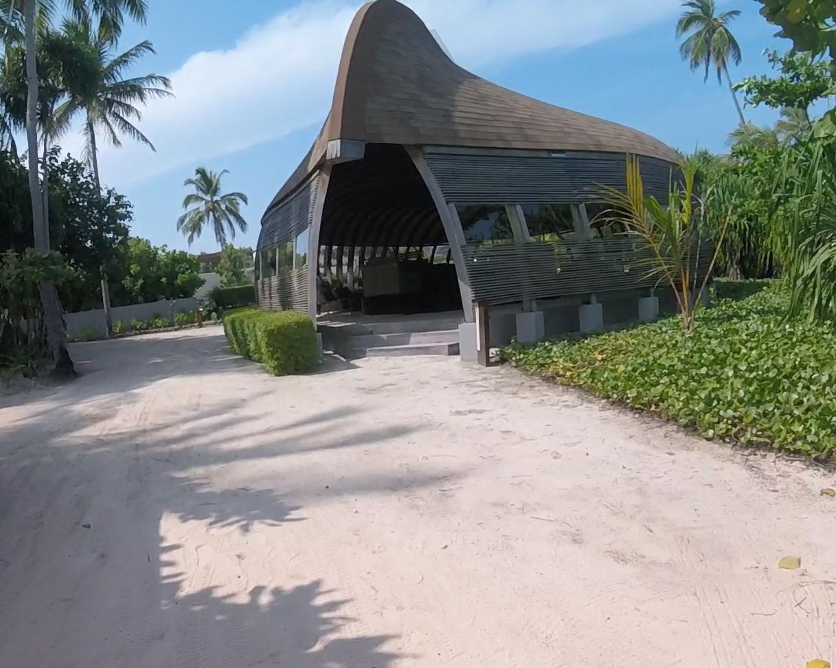 A distinctive building with a curved roof stands on a sandy path surrounded by tropical trees at the Park Hyatt Maldives.