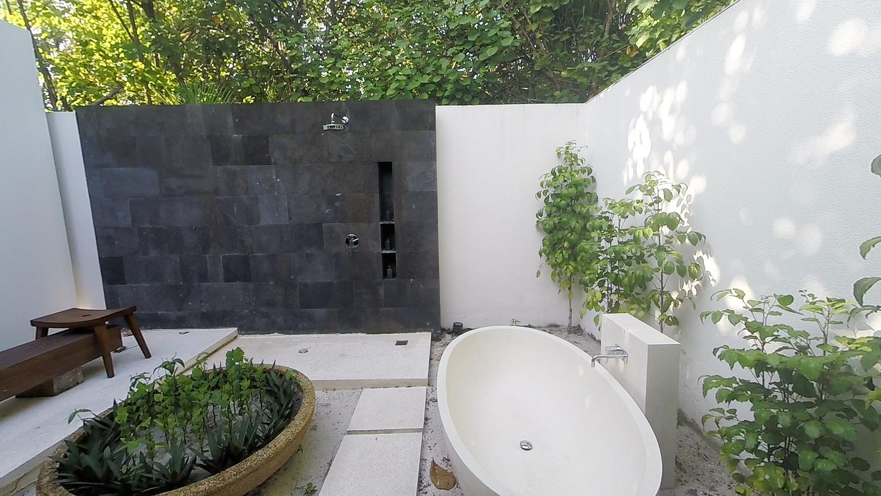 Outdoor bathroom with a black tiled shower wall, white freestanding bathtub, and green plants.