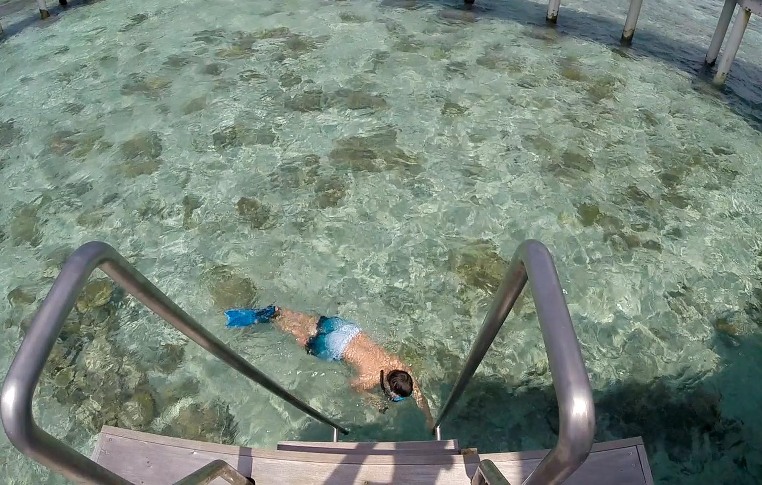 A person snorkels in clear turquoise water below steps from a water villa.