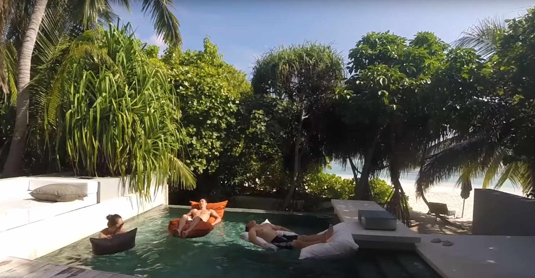 Three people relax on floats in a pool surrounded by tropical plants, with a beach and ocean visible beyond.