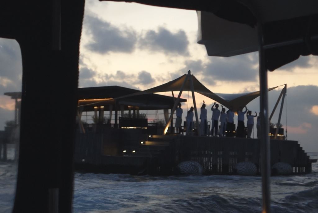 Silhouetted resort staff wave goodbye from a platform over the ocean at sunset.