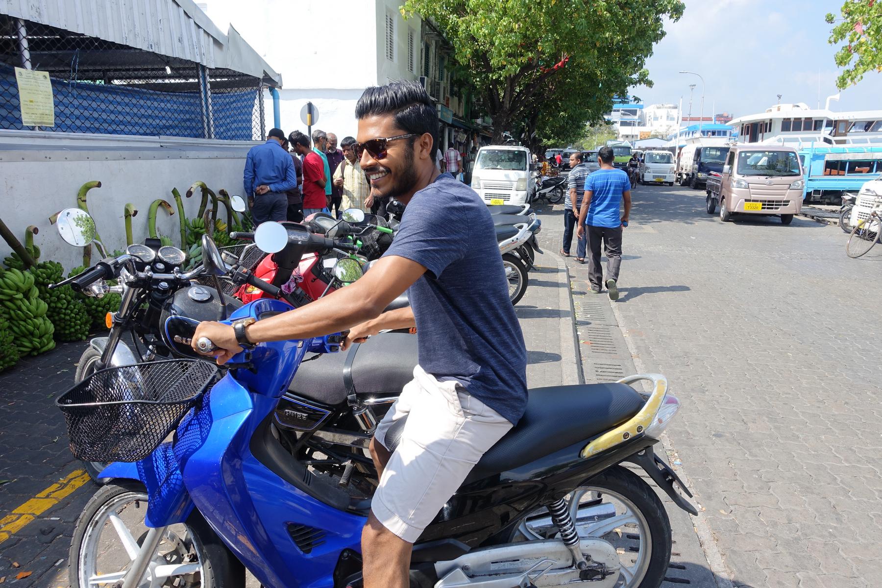 A smiling man sits on a blue scooter in a bustling street scene in Male, Maldives.
