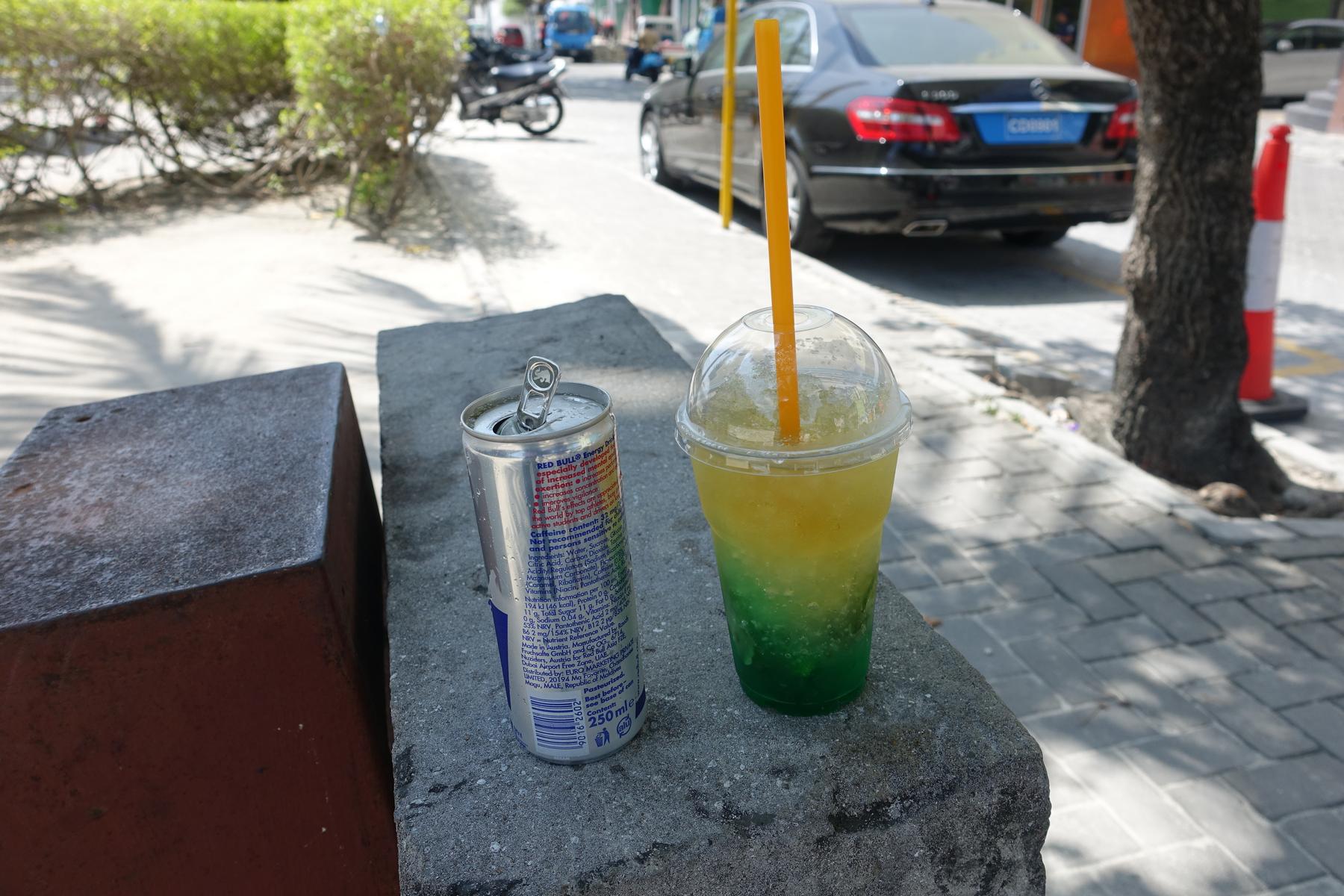 A can of Red Bull and a layered green and yellow slushy drink sit on a concrete ledge next to a street in Male.
