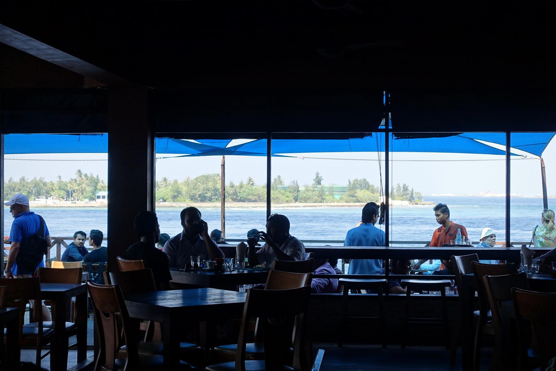 Silhouetted people sit inside a restaurant with large windows overlooking the ocean and a tropical island.