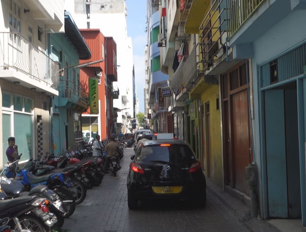 A narrow street in Male lined with colorful buildings, many parked motorbikes, and a black car driving away.
