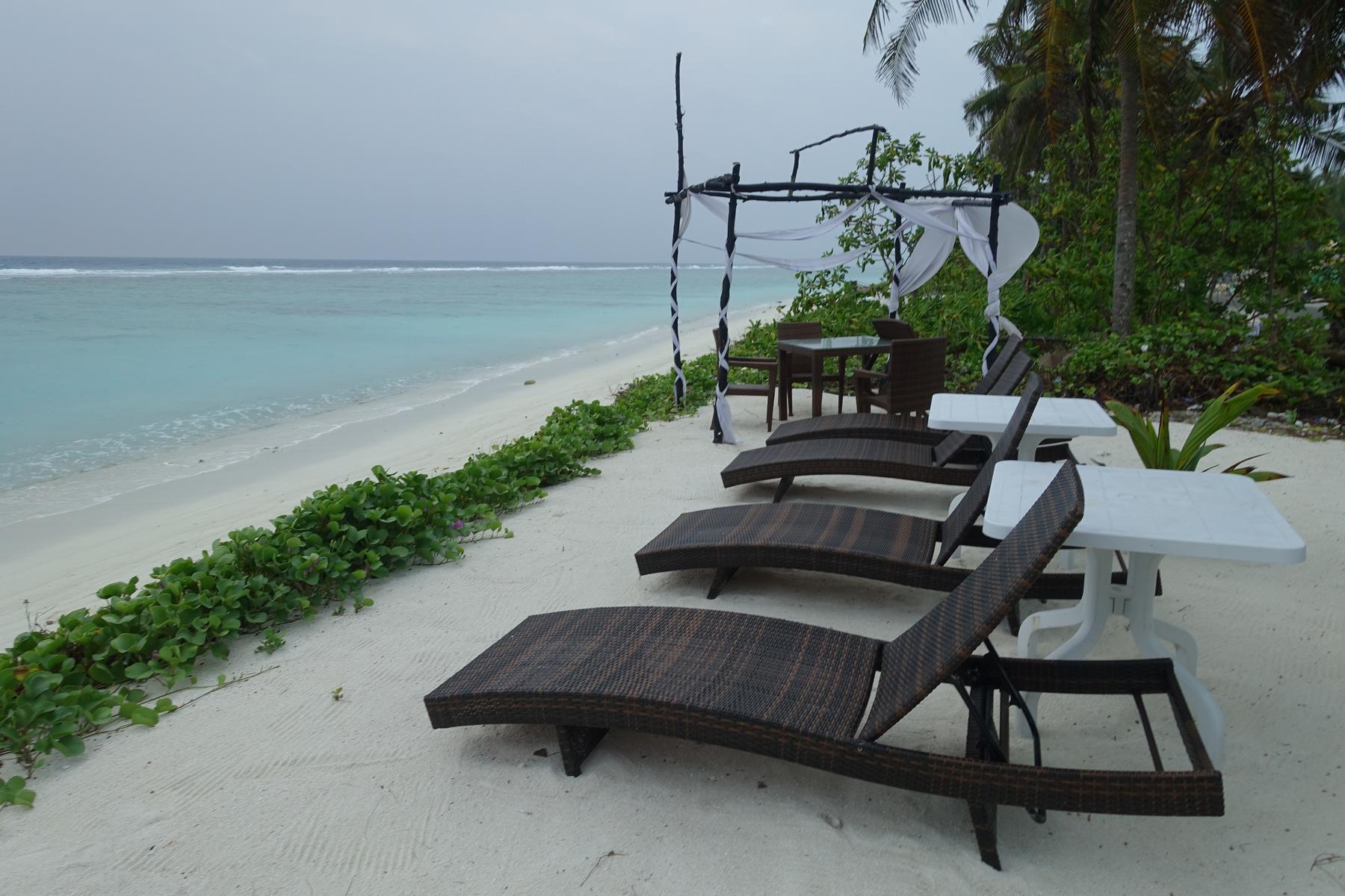 Beach loungers and a shaded dining area on white sand by the turquoise ocean on an overcast day.