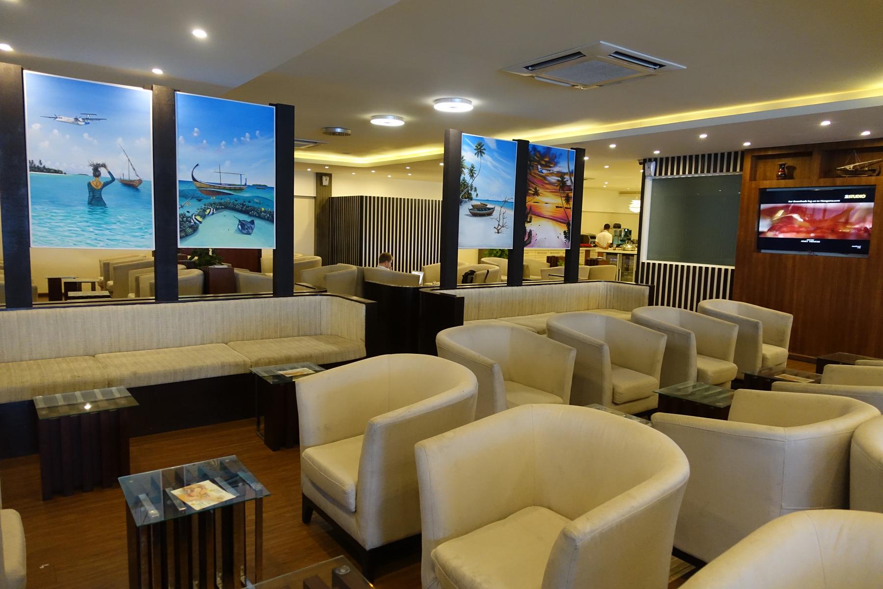 A bright, modern lounge with rows of cream seating, wooden accents, and large wall art depicting Maldivian island scenes.