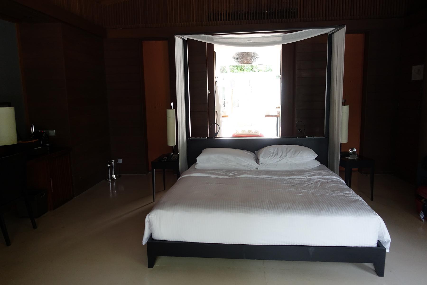 A dark wooden bedroom features a canopy bed with white linens, facing a brightly lit open doorway.