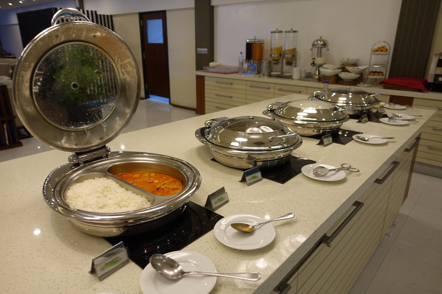 A buffet counter features several chafing dishes, with an open one revealing white rice and an orange curry.