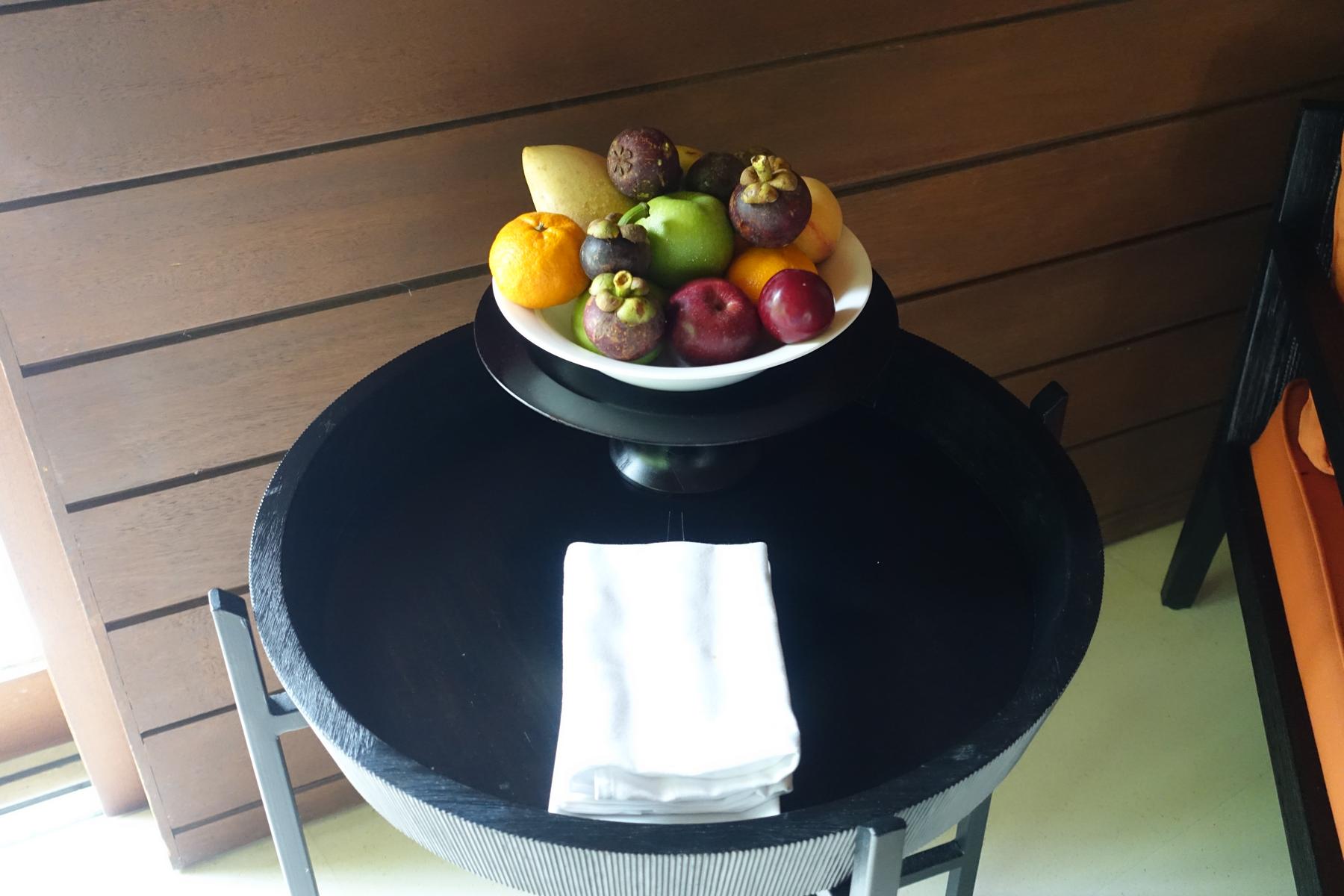 A white bowl overflowing with colorful fruits, including mangosteens, oranges, and apples, sits on a black side table with a folded white napkin.
