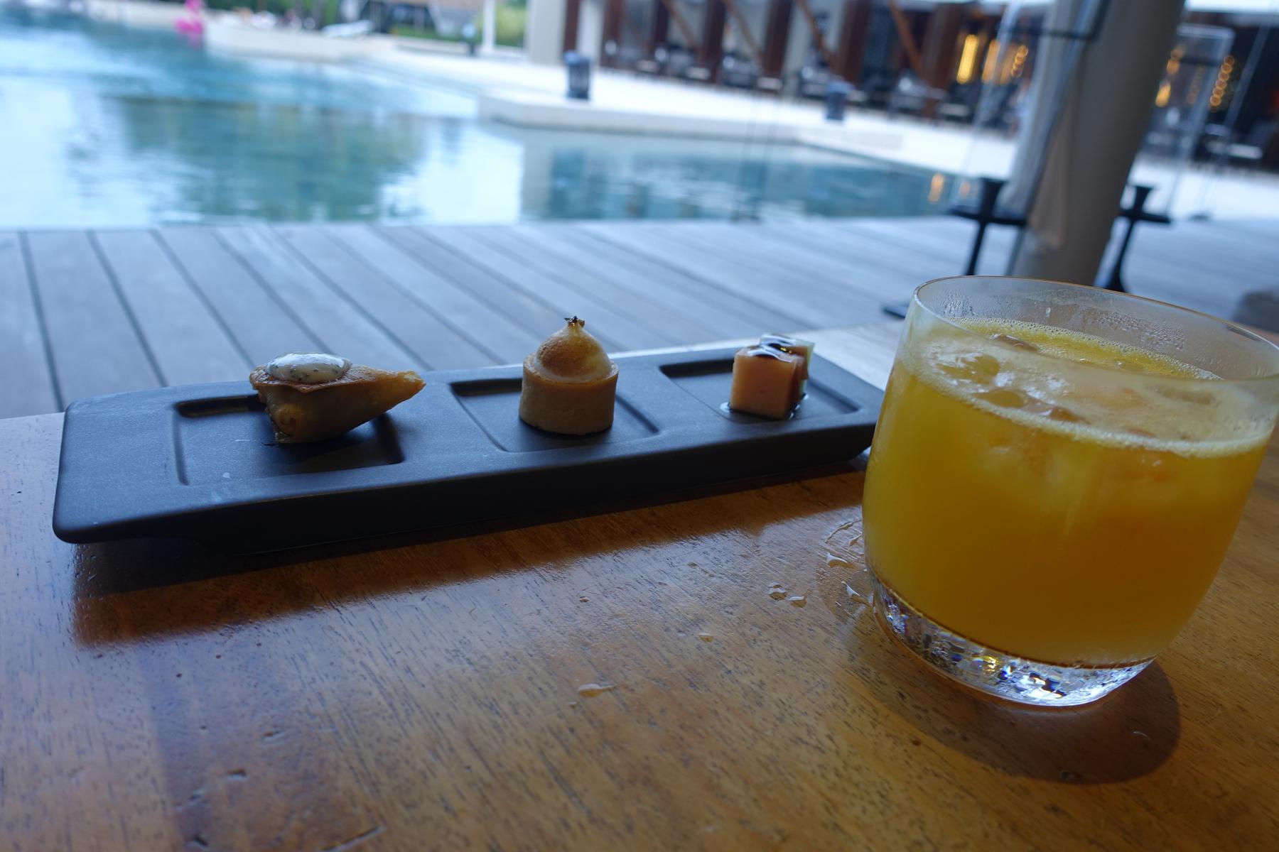 Three small appetizers and a glass of orange juice sit on a wooden table next to a swimming pool.