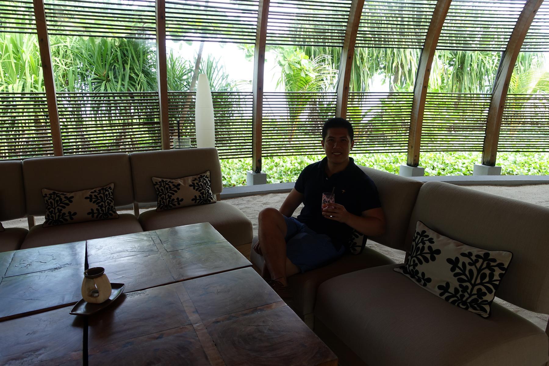 A man smiles on a sofa with a drink in a resort lounge overlooking lush tropical greenery.