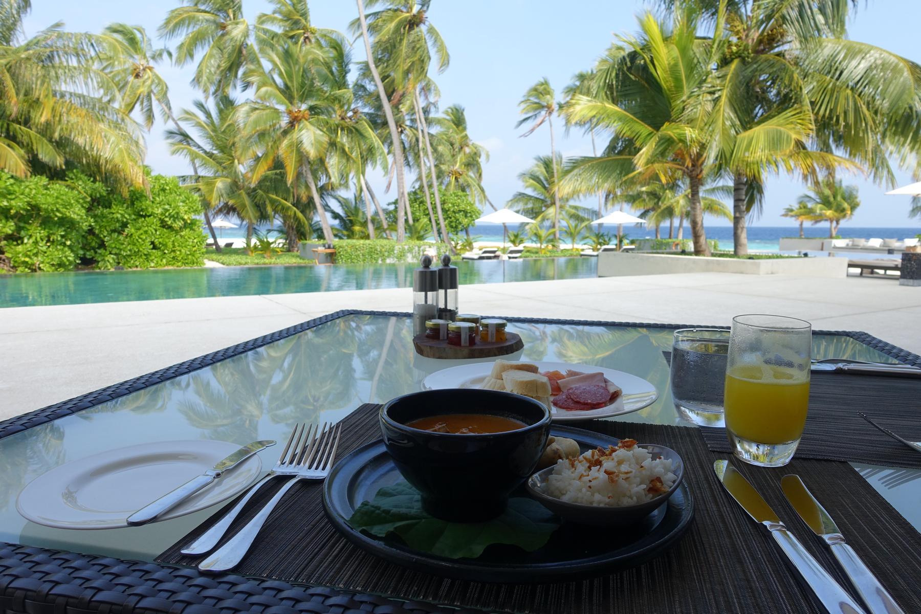 Breakfast is served on a glass table overlooking an infinity pool, palm trees, and the ocean.