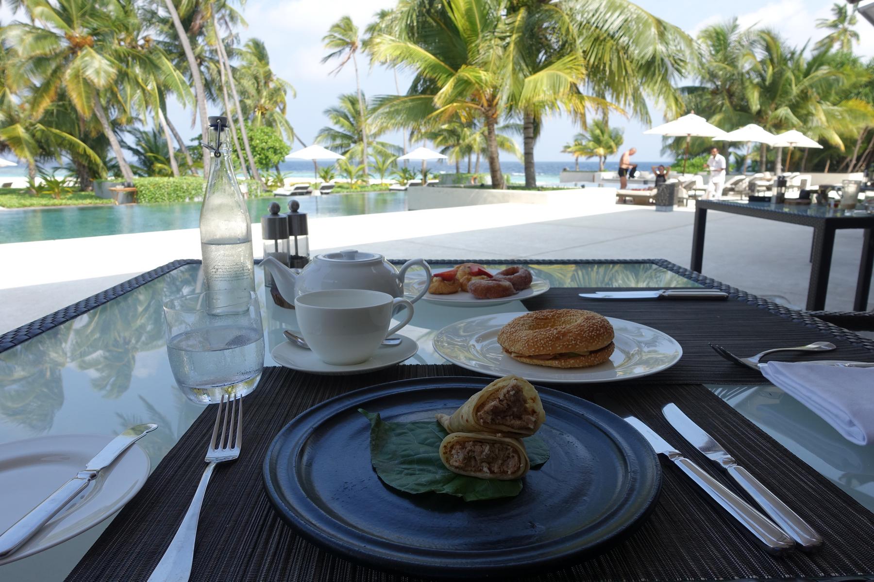 An outdoor breakfast of wraps, a bagel, pastries, and tea is set on a glass table overlooking a resort pool, palm trees, and the ocean.
