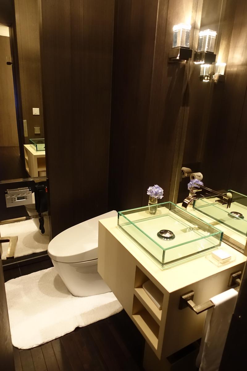 A modern hotel bathroom featuring dark wood walls, a white toilet, and a glass basin on a light vanity with a mirror.