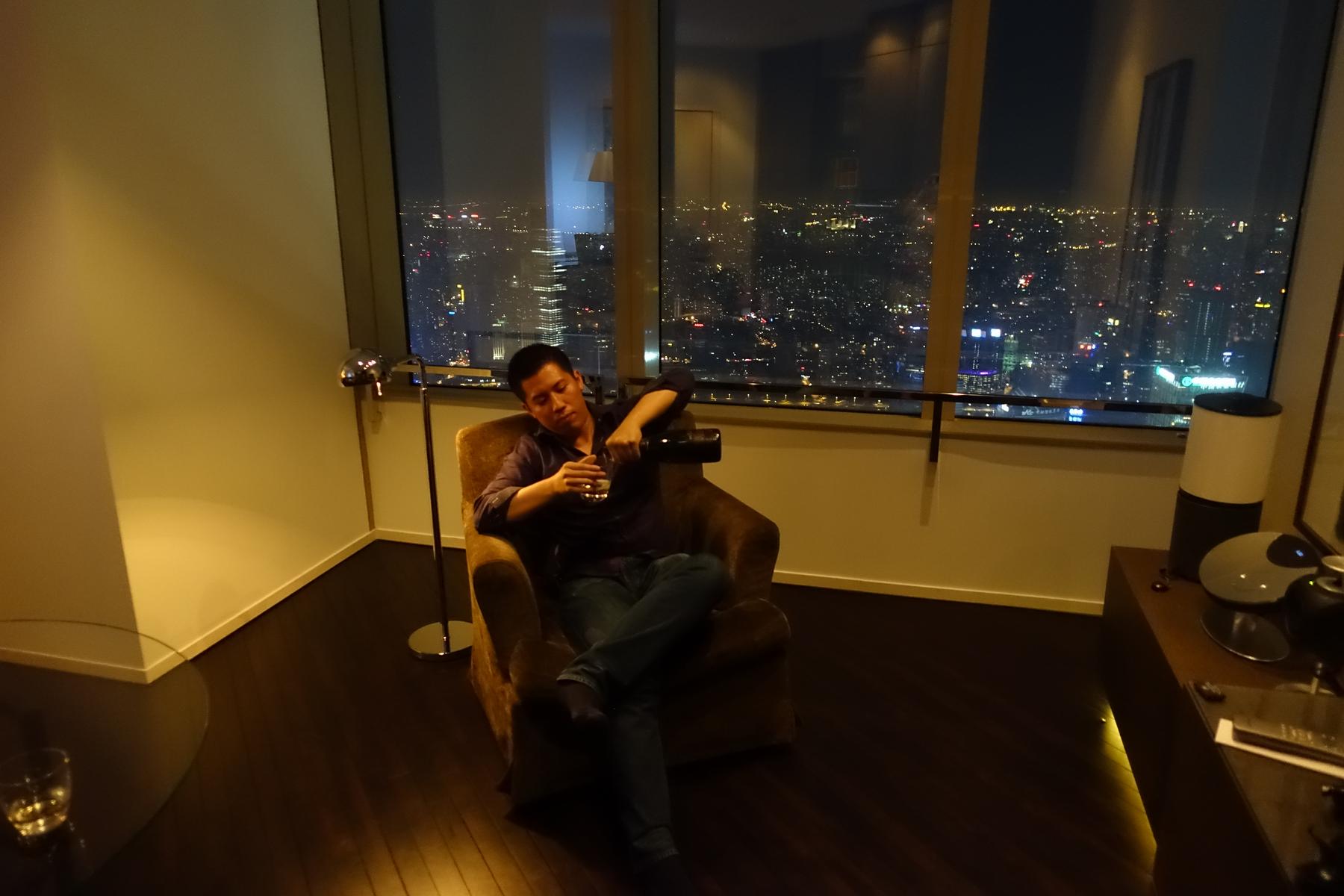 A man sits in an armchair pouring a drink, enjoying a panoramic city lights view from a high-rise suite at night.