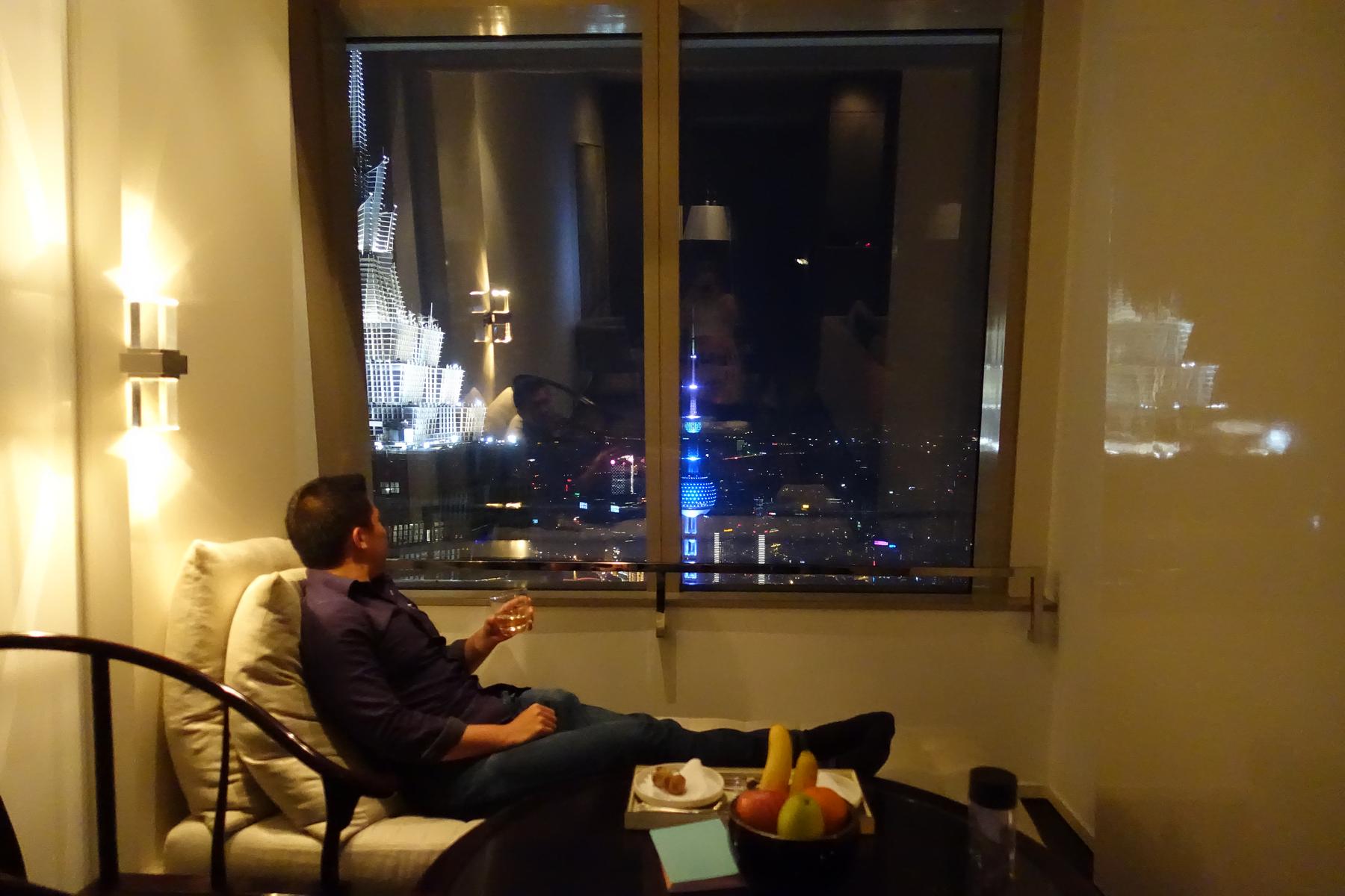 A man relaxes on a lounge chair, looking out a large window at the illuminated Shanghai skyline at night.