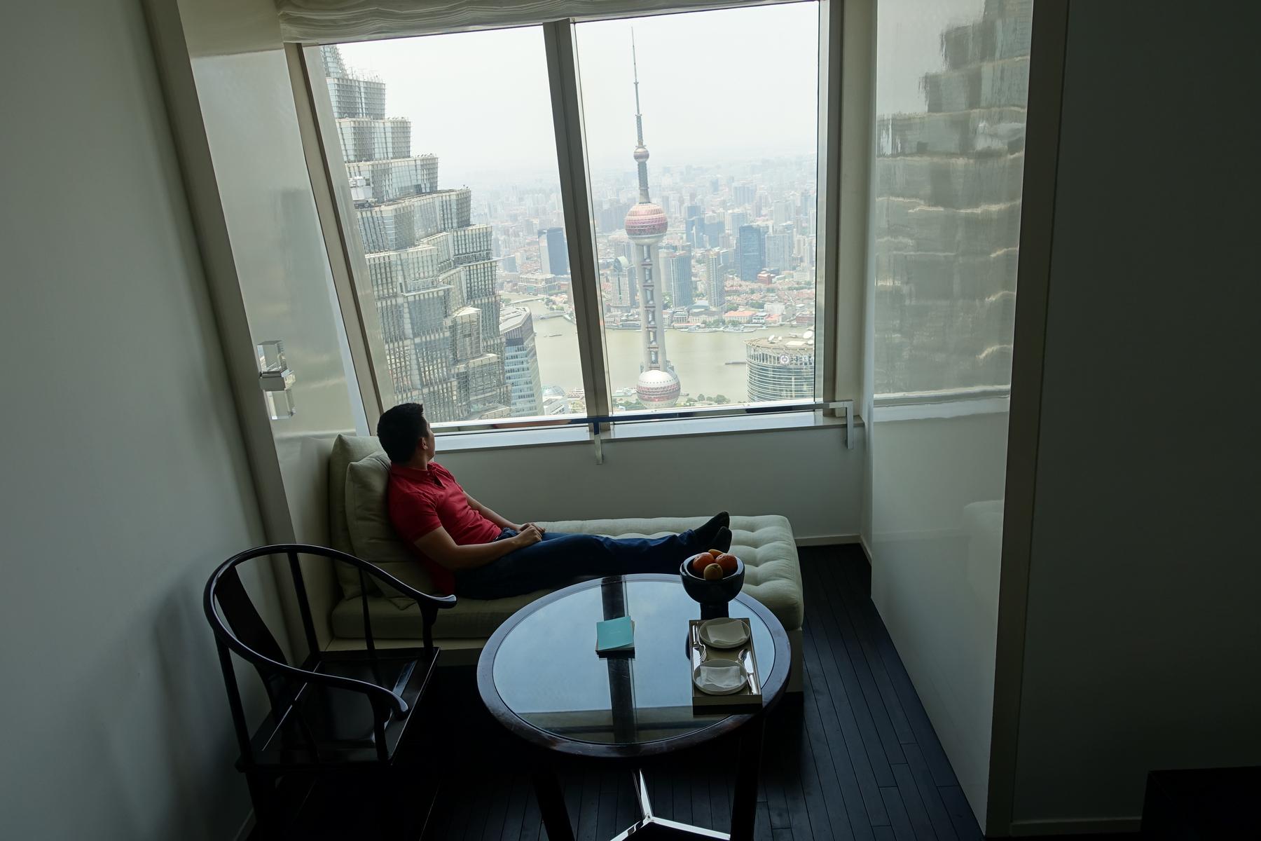 A man reclines on a window-side daybed, admiring the Shanghai skyline including the Oriental Pearl TV Tower.
