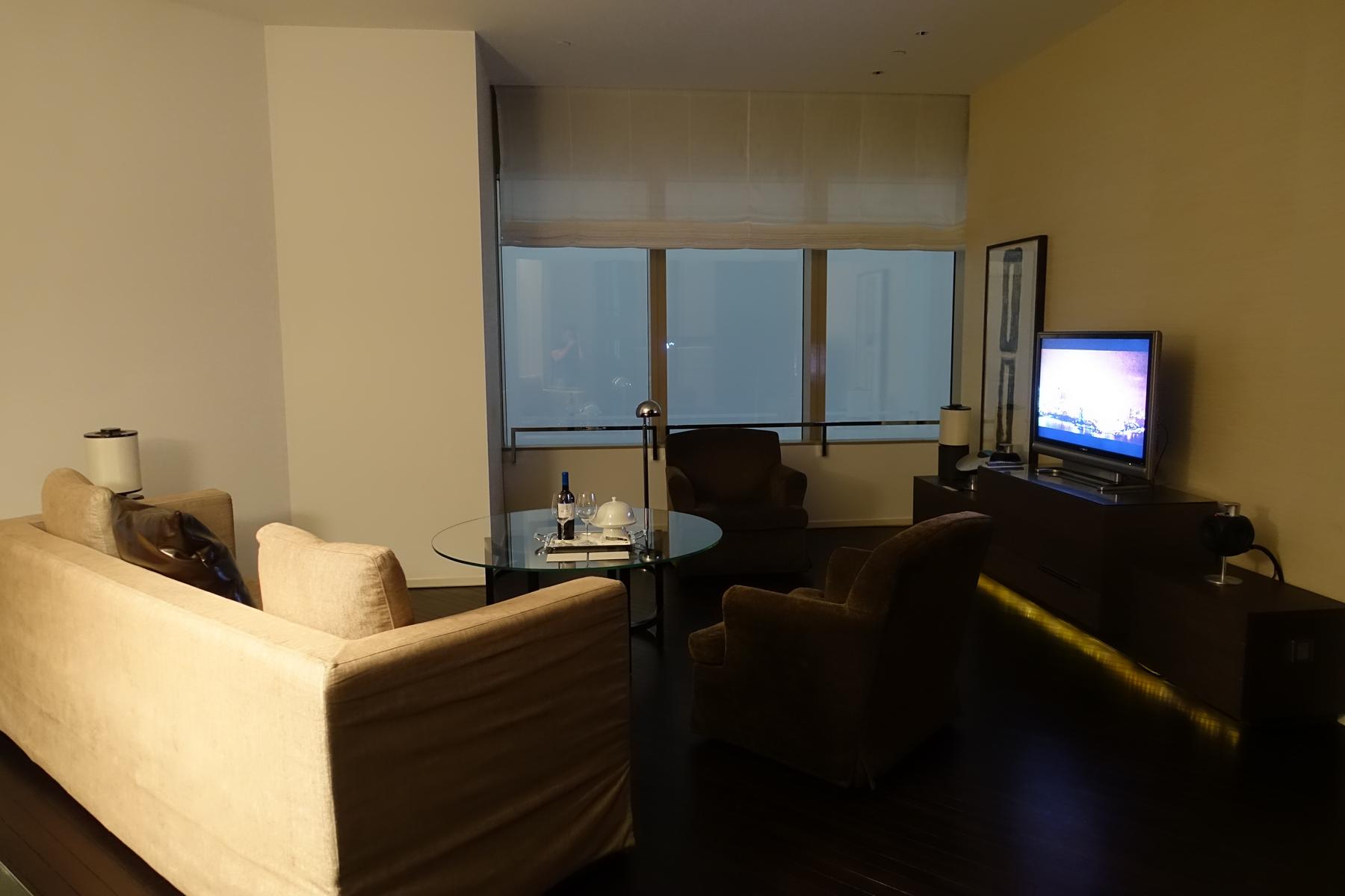 A Park Hyatt Shanghai Bund View Suite living room with a sofa, armchairs, TV, and glass coffee table, overlooking a city night scene through large windows.