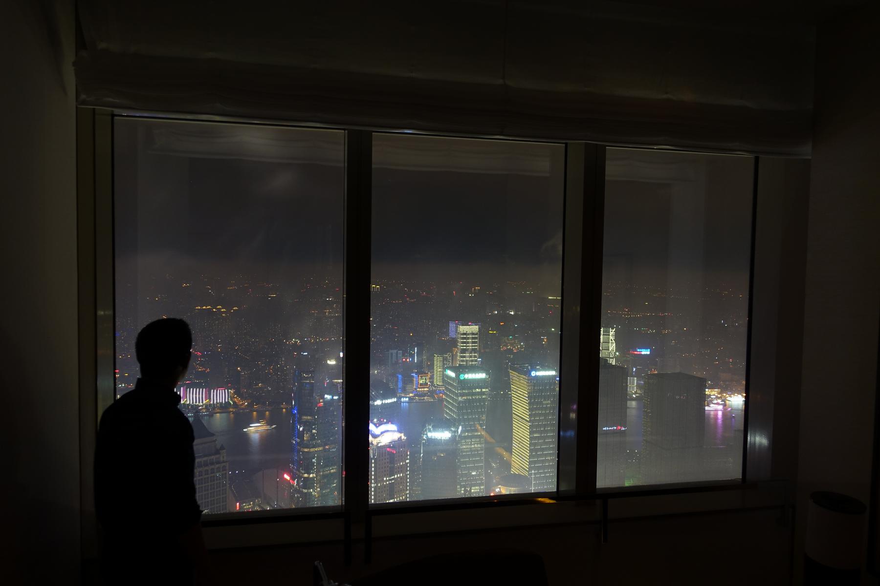 Silhouette of a person looking out a window at the illuminated Shanghai skyline and the Bund at night.