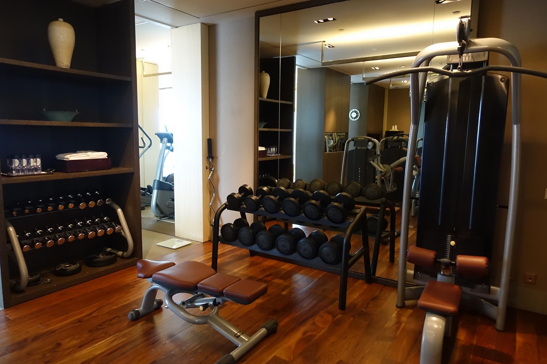 A well-equipped hotel gym with a dumbbell rack, weight bench, and exercise machines, featuring a mirrored wall and wooden floor.