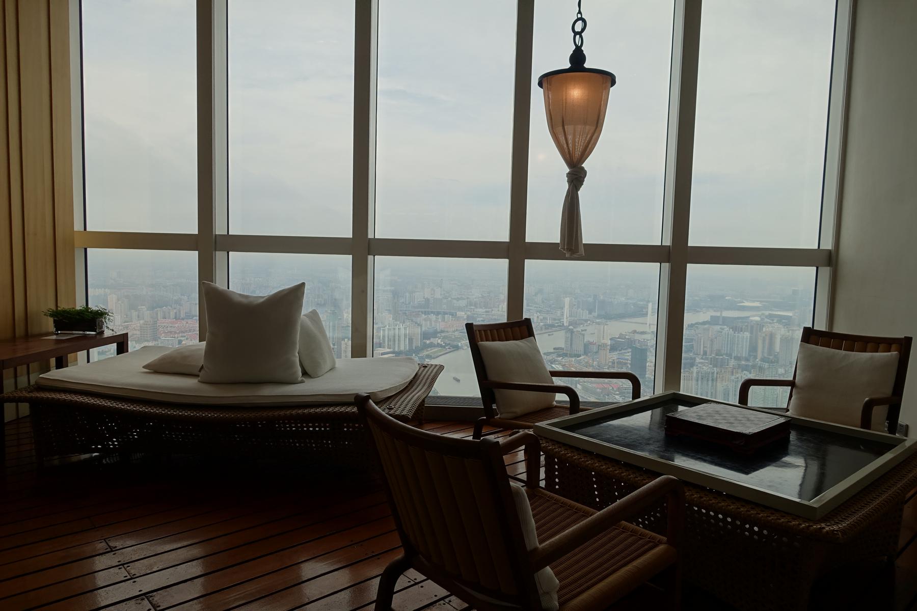 A lounge with a daybed, chairs, and a table, overlooking the Shanghai skyline through large windows.