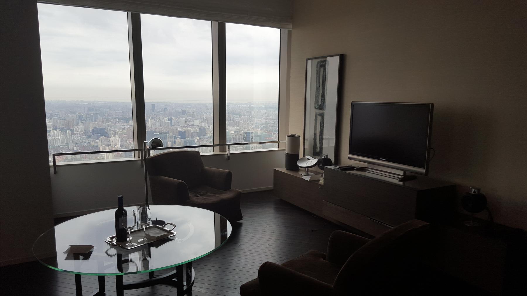 Hotel suite living room with a city view, TV, and wine on a glass table.