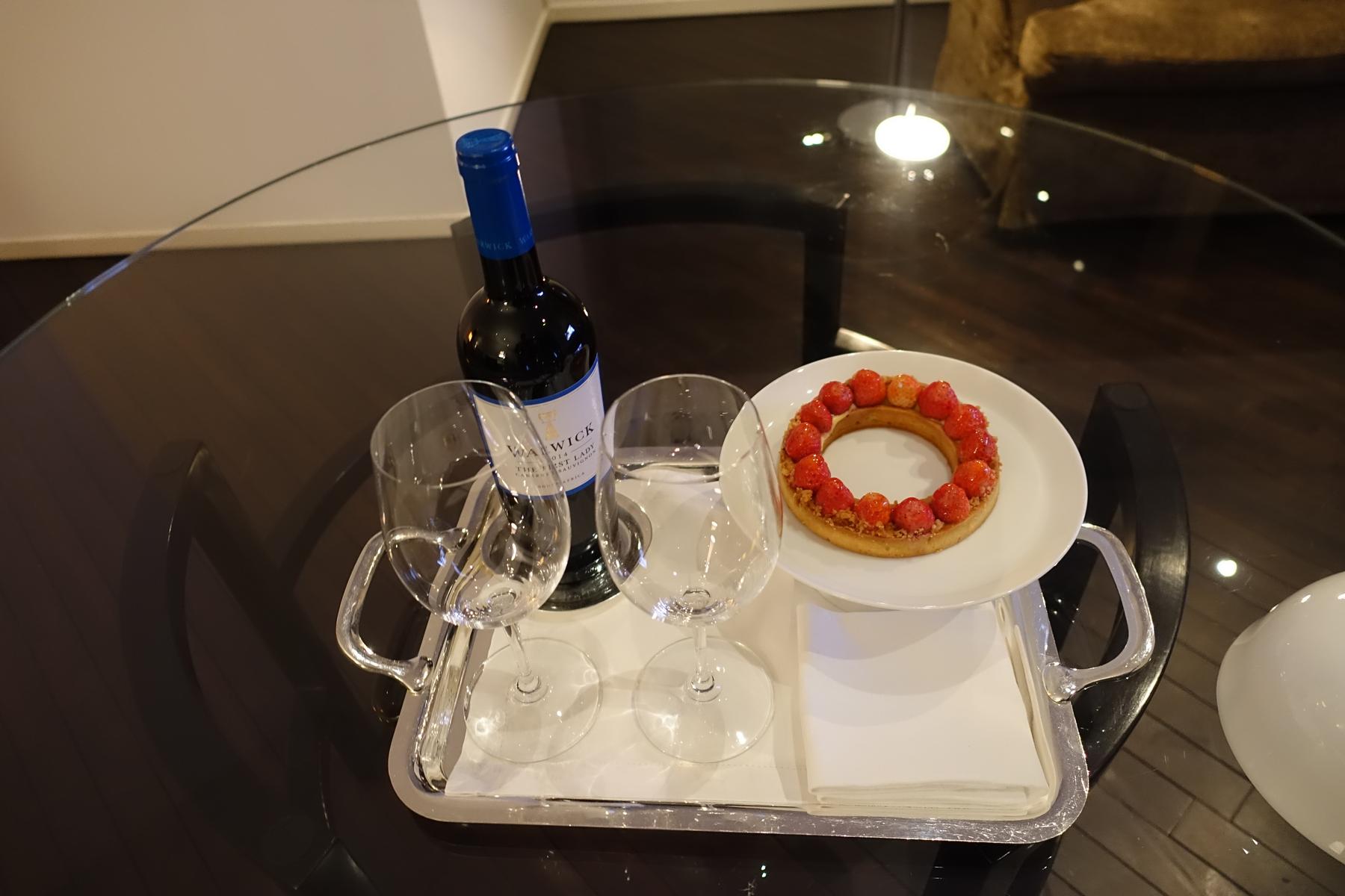 A silver tray on a glass table holds a bottle of wine, two glasses, and a strawberry tart.
