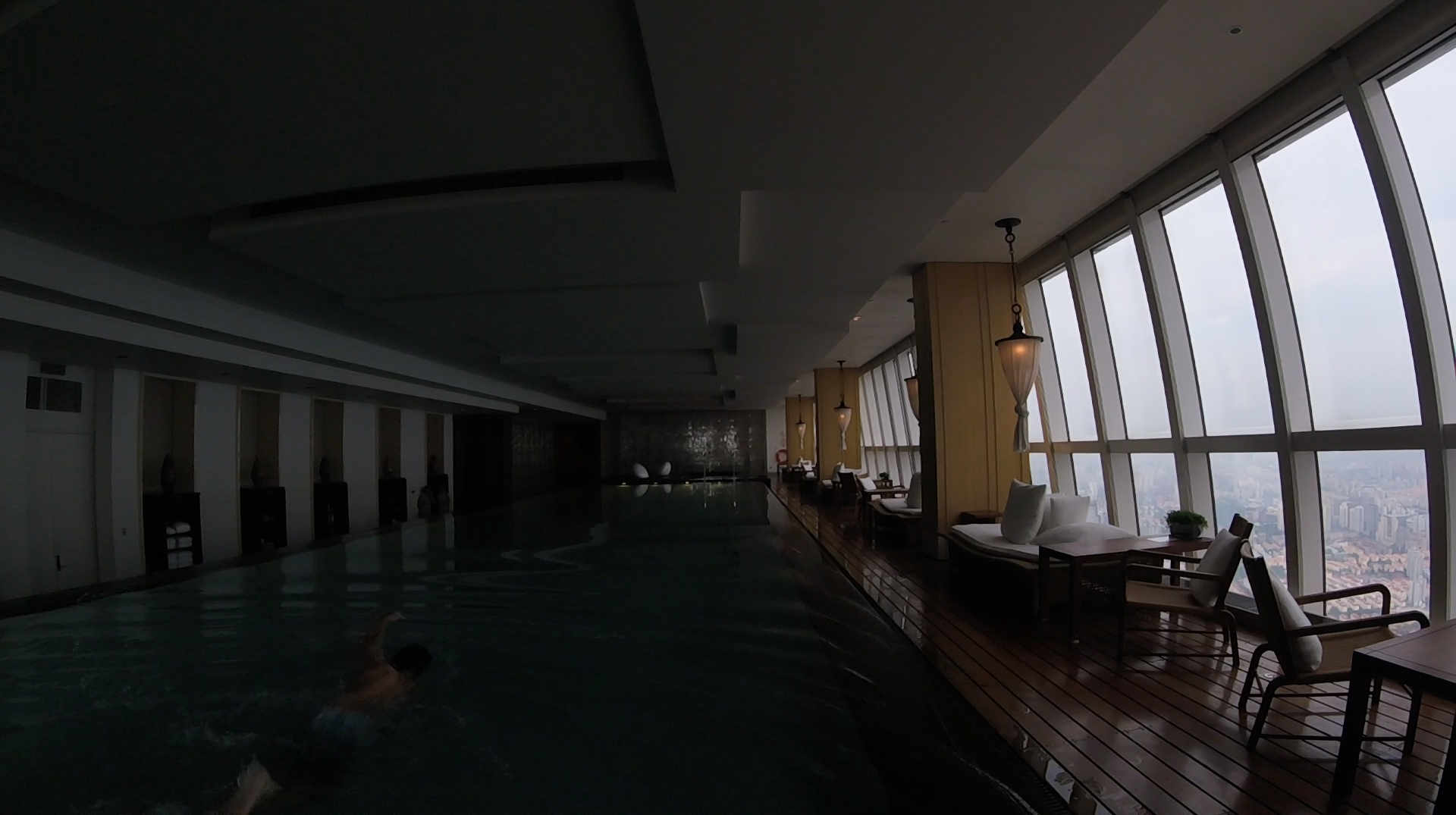 A long indoor swimming pool with a swimmer and a lounge area next to tall windows with a city view.