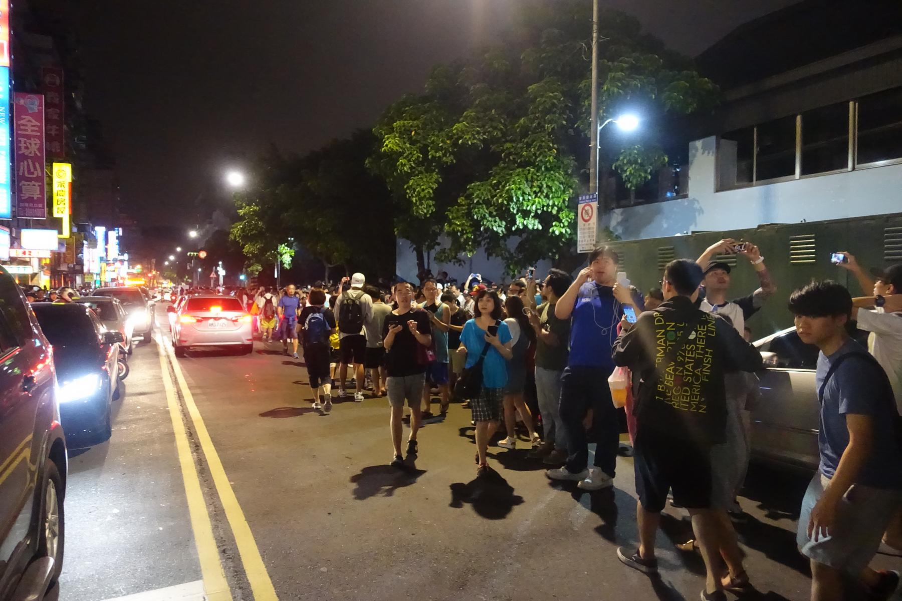 A large crowd of people, many holding phones, fills a street in Taiwan at night.