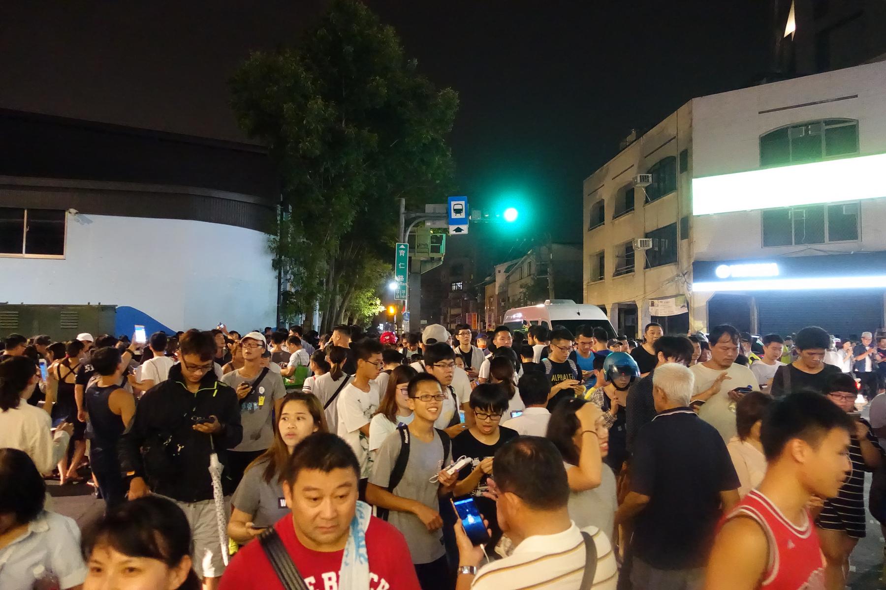 A dense crowd of people, many looking at their phones, fills a street in Taiwan at night.