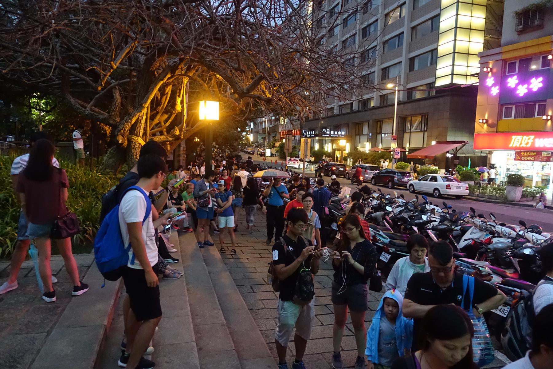 A dense crowd of people on a Taiwan street at dusk, most looking at phones near parked scooters.