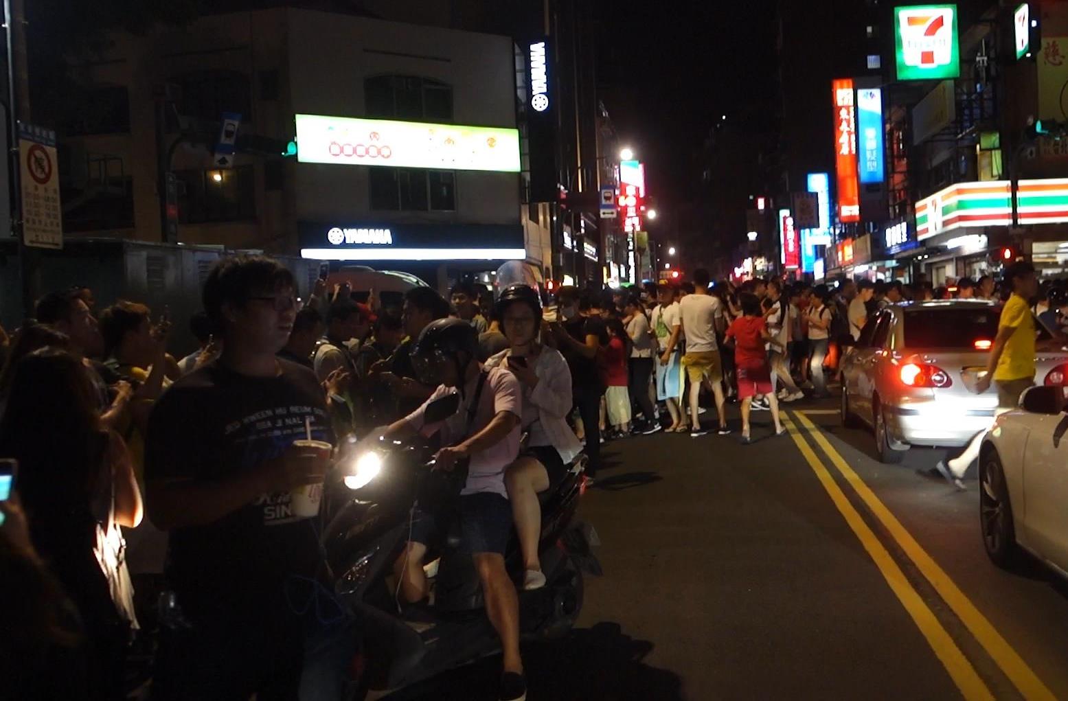 A crowded street at night in Taiwan, filled with people and scooters, suggesting a Pokemon Go gathering.
