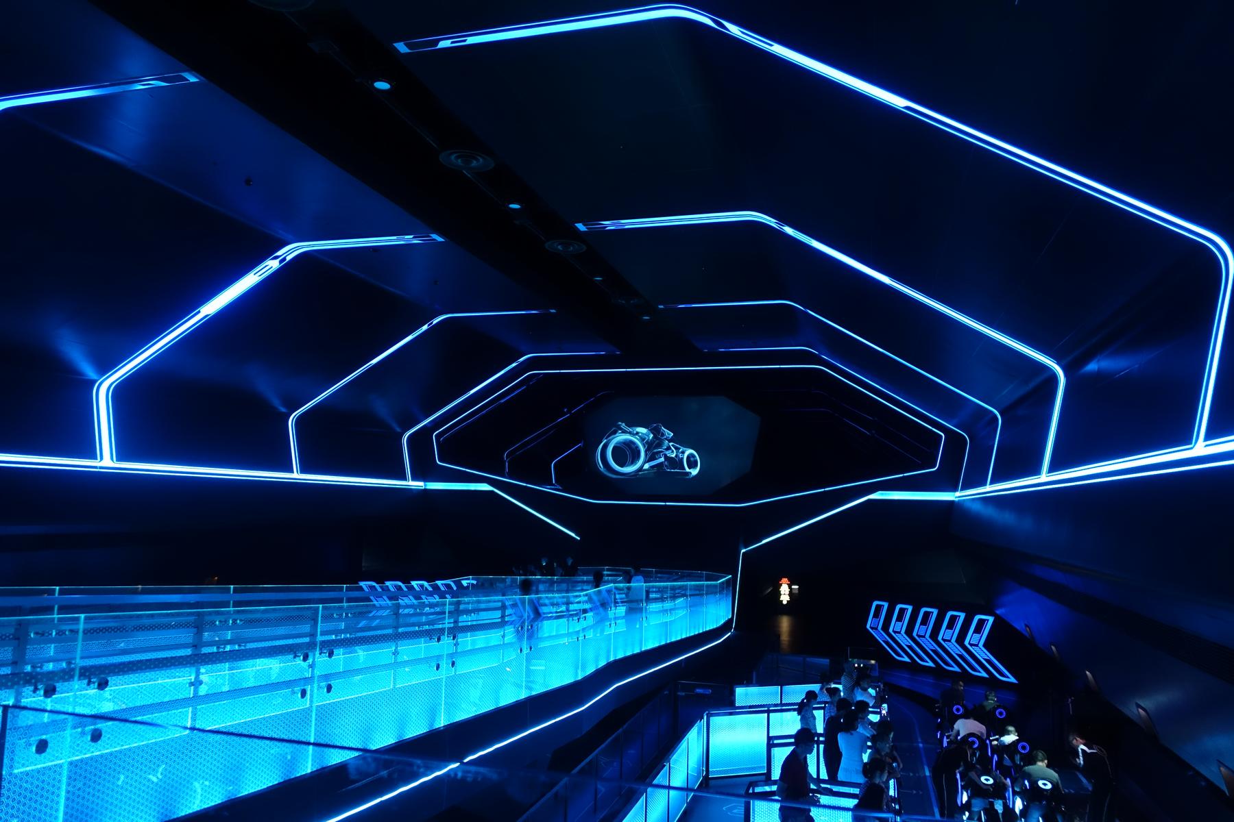 The futuristic, dark blue TRON Lightcycle ride loading area features glowing neon outlines, a projected Lightcycle image, and guests preparing to board.
