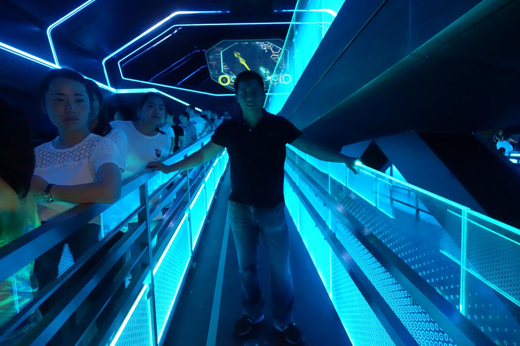 A man smiles and poses in a brightly lit blue, futuristic TRON-themed ride queue.