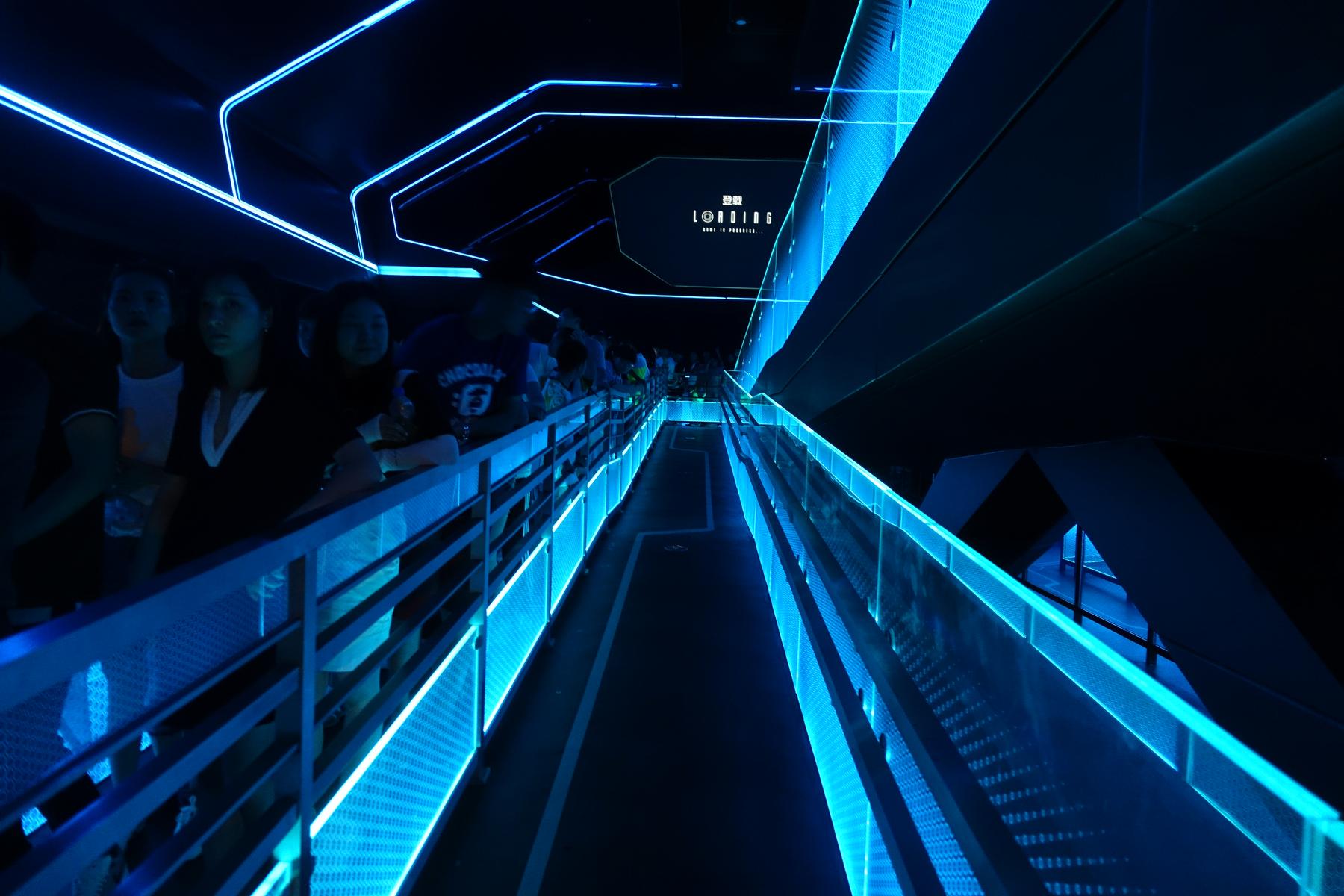 A dark, futuristic queue line glowing with neon blue lights, where people wait beneath a 'LOADING' screen.
