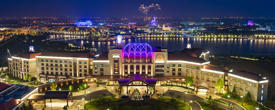 Aerial night view of Shanghai Disneyland Hotel with the illuminated park and fireworks in the background.
