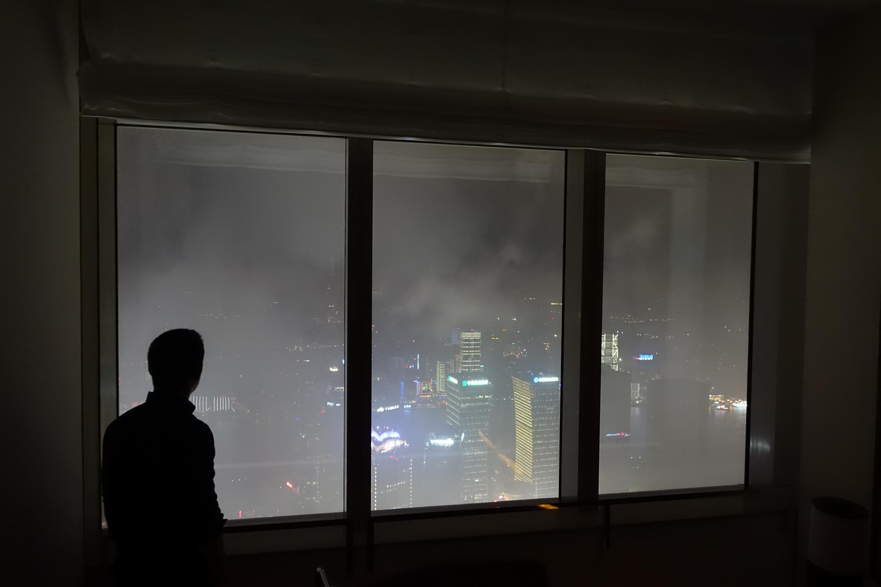 A silhouetted person looks out a tall window at a cloudy night city view.