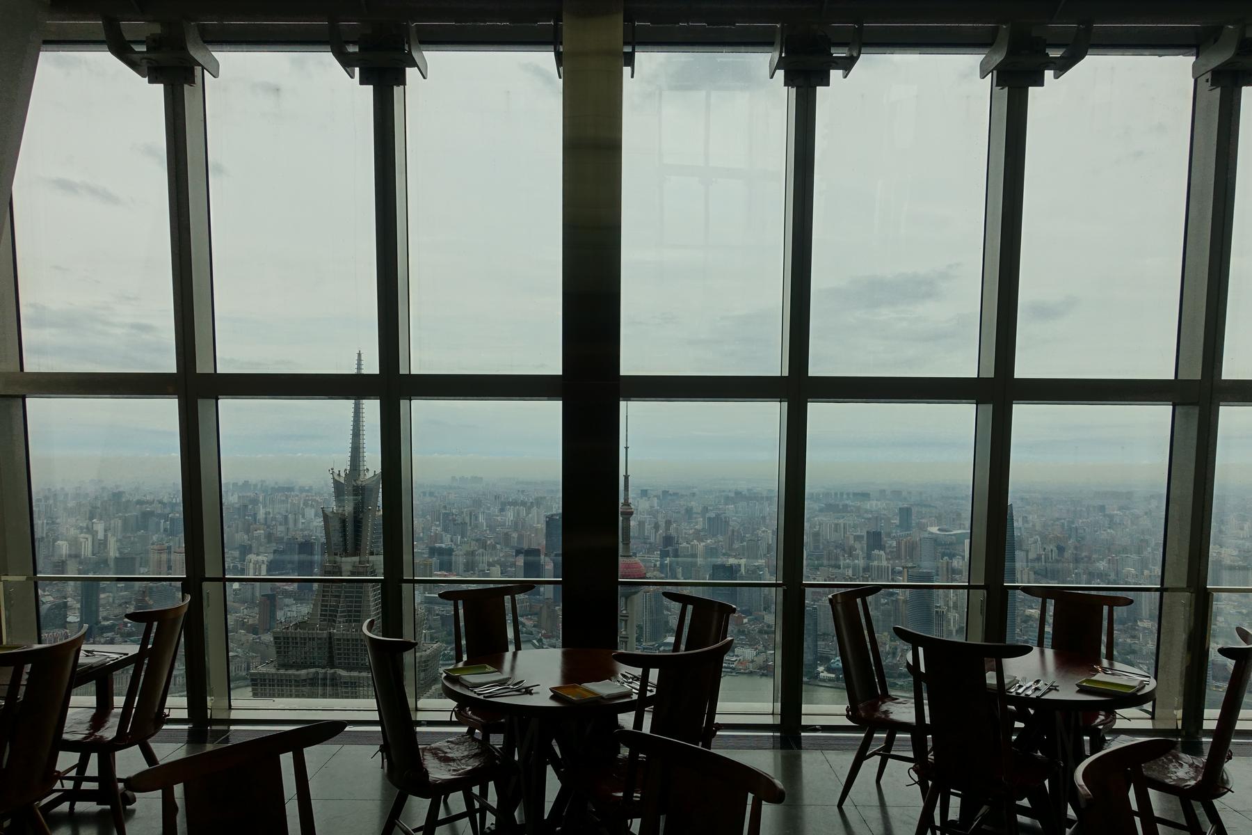 High-rise restaurant with dark wooden tables and chairs overlooking a vast Shanghai cityscape.