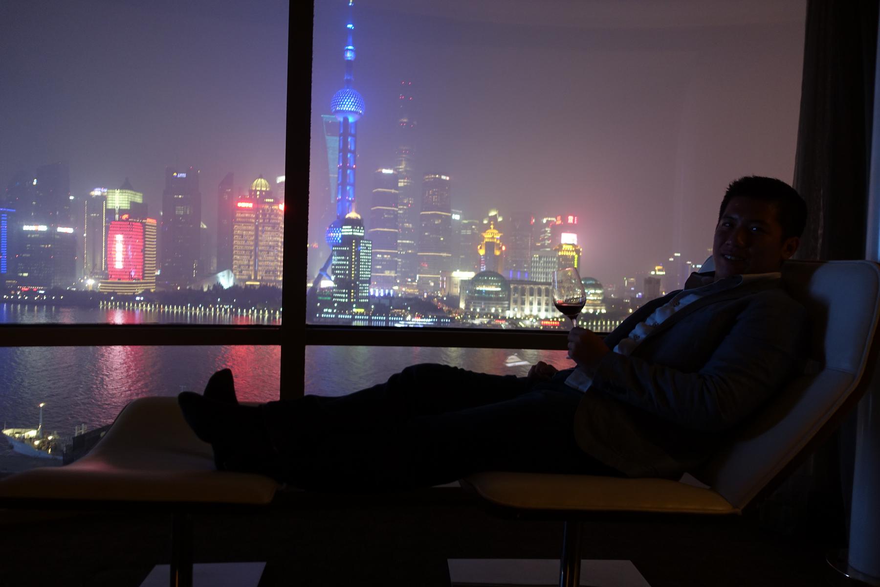 Man relaxes with wine, overlooking the dazzling Shanghai night skyline from a high-rise window.