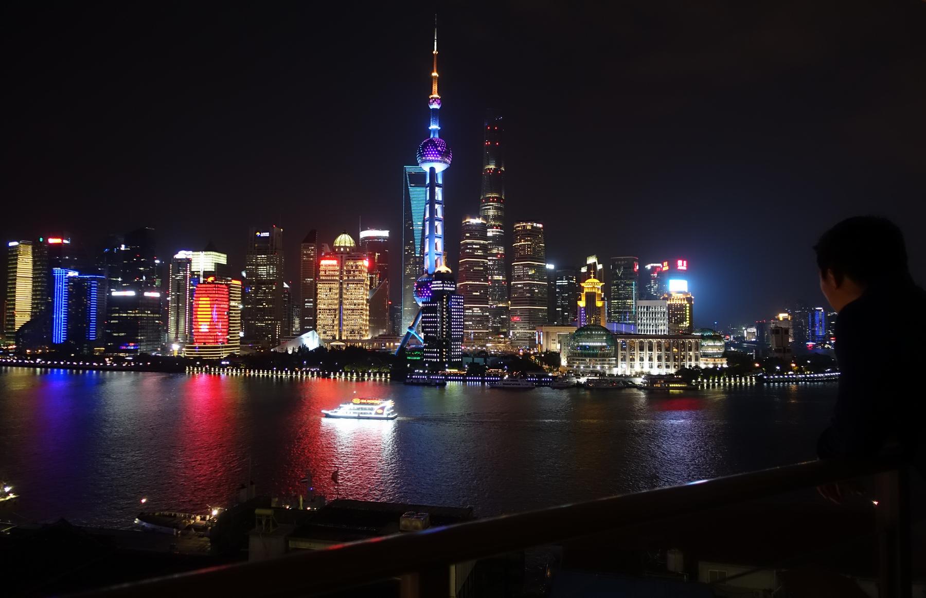 Person overlooking the illuminated Shanghai skyline and Huangpu River at night.