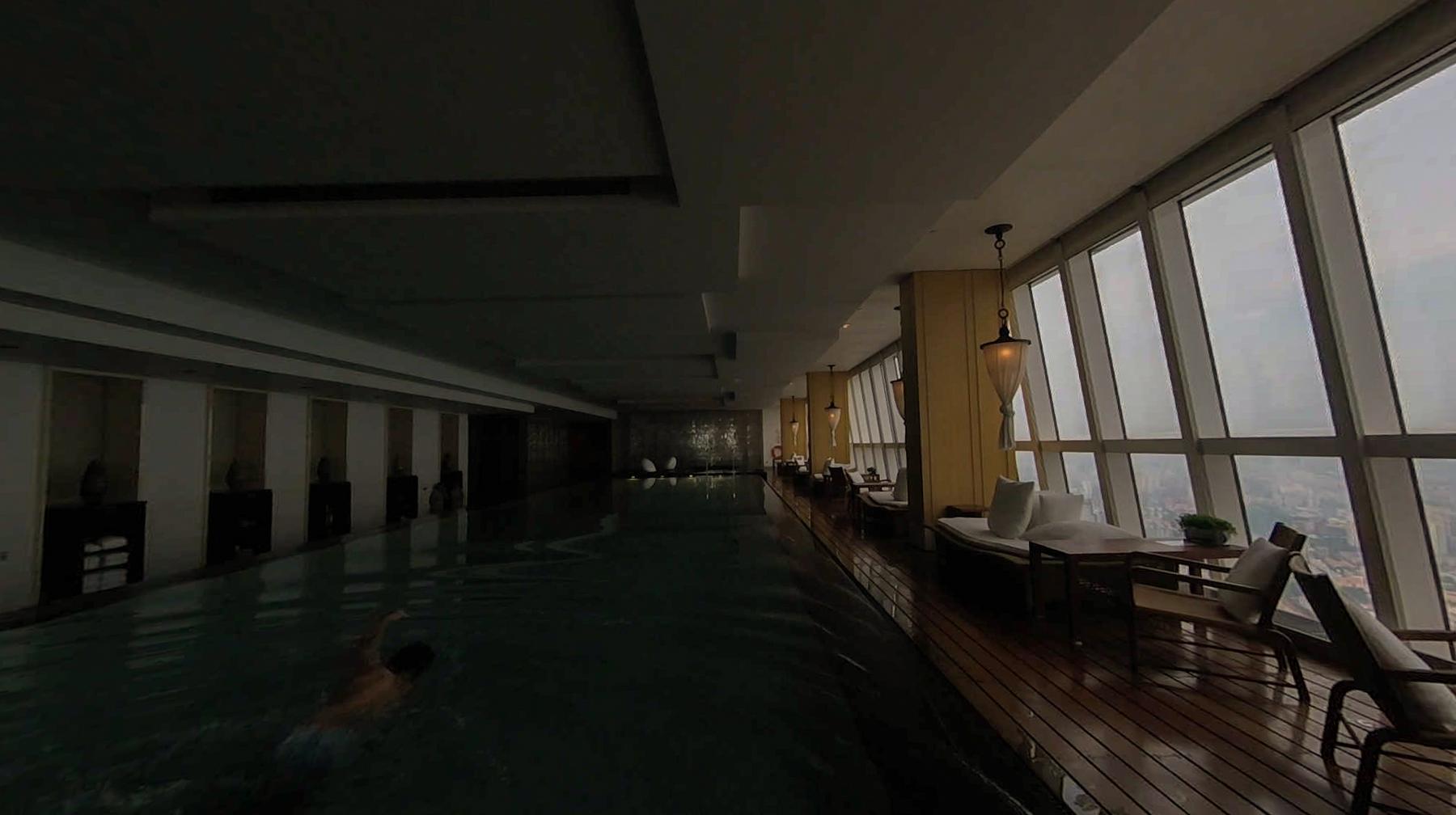 A person swims in a high-rise indoor pool with a lounge area next to large windows overlooking a city.