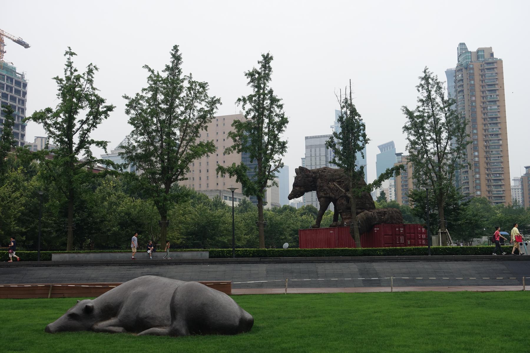 A modern city park features two large bear sculptures, one lying on grass and another standing on red shipping containers, with tall buildings in the background.