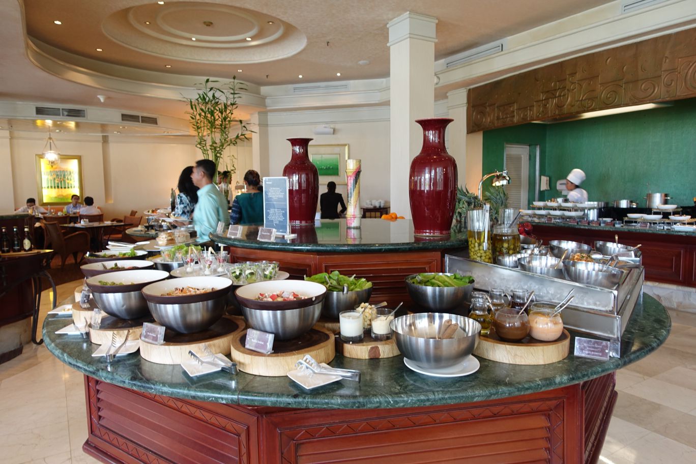 A bright restaurant features a circular buffet table laden with salads and other dishes, with guests and a chef in the background.