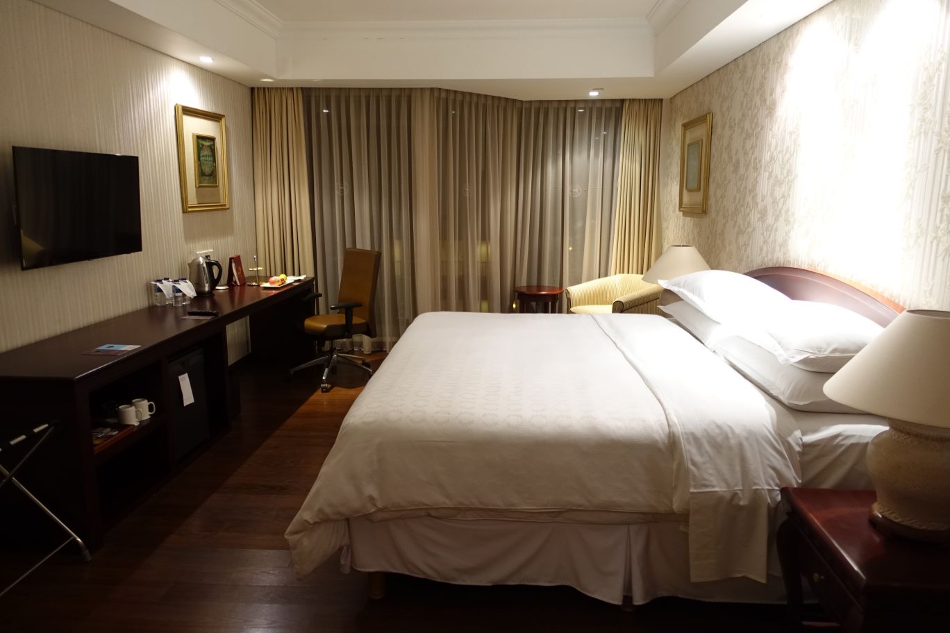 A well-lit hotel room features a large bed, a desk with a TV, and floor-to-ceiling curtains.
