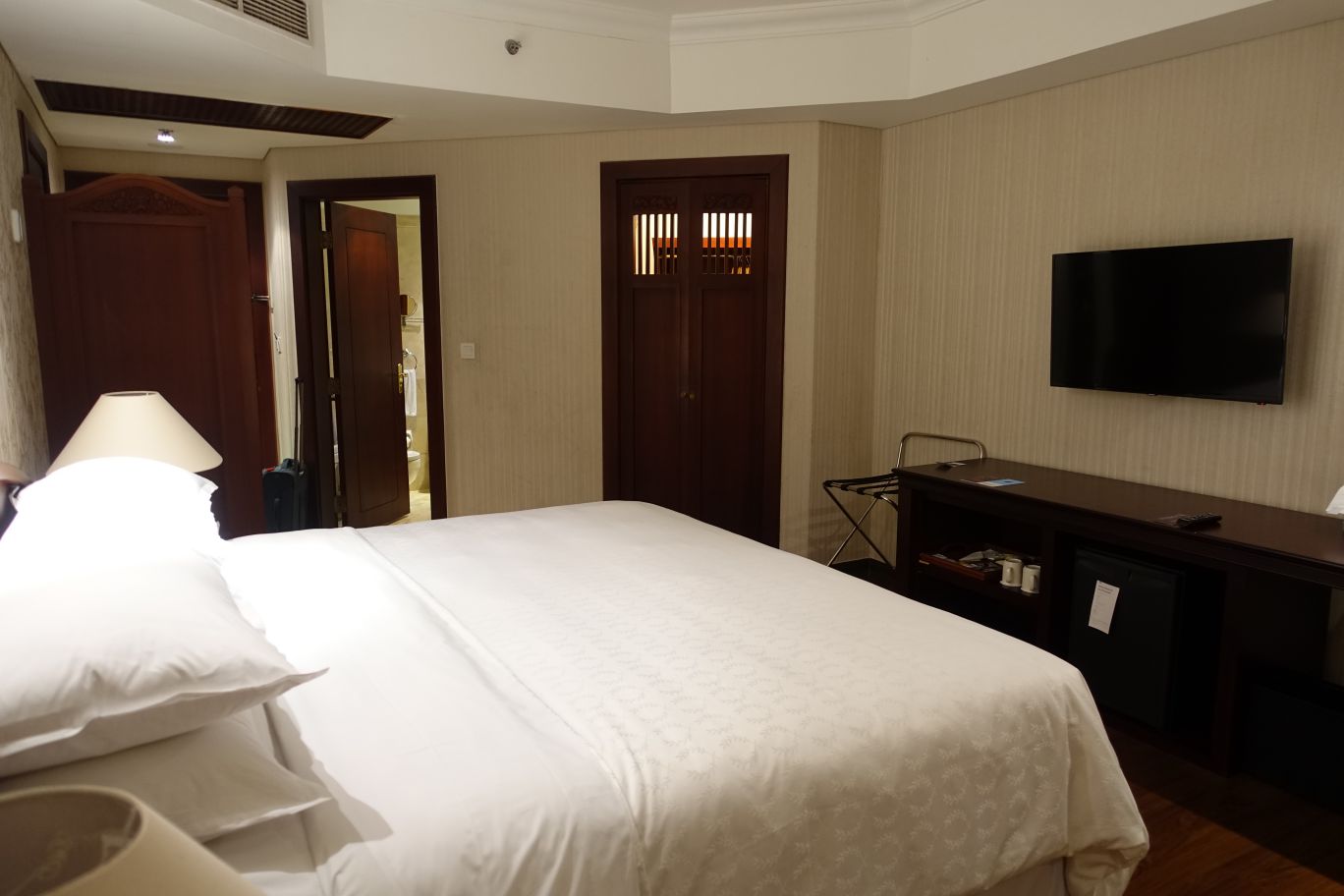 A hotel room with a large white bed, dark wood furniture, and a wall-mounted TV.
