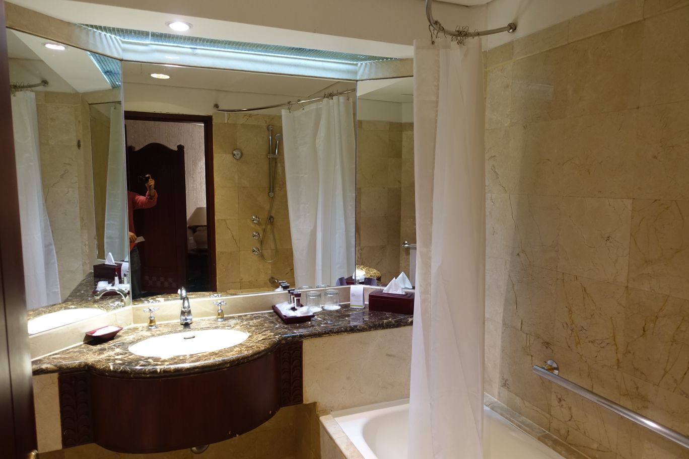 A clean hotel bathroom with light marble walls, a dark wood vanity with a white sink, and a bathtub with a white shower curtain.