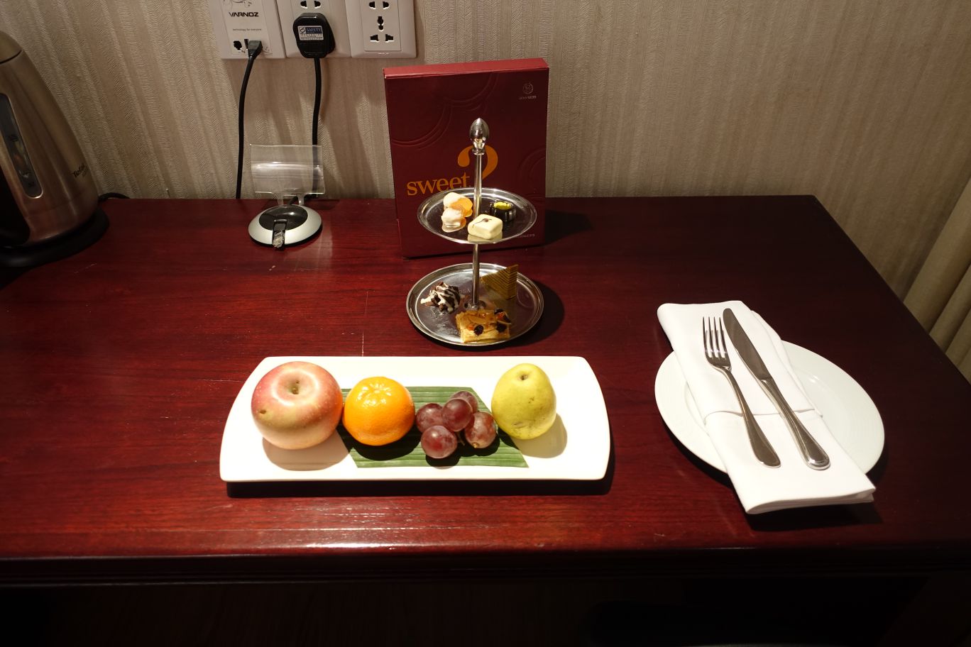 Hotel welcome amenities including a fruit platter, a stand with sweets, and a place setting on a wooden table.